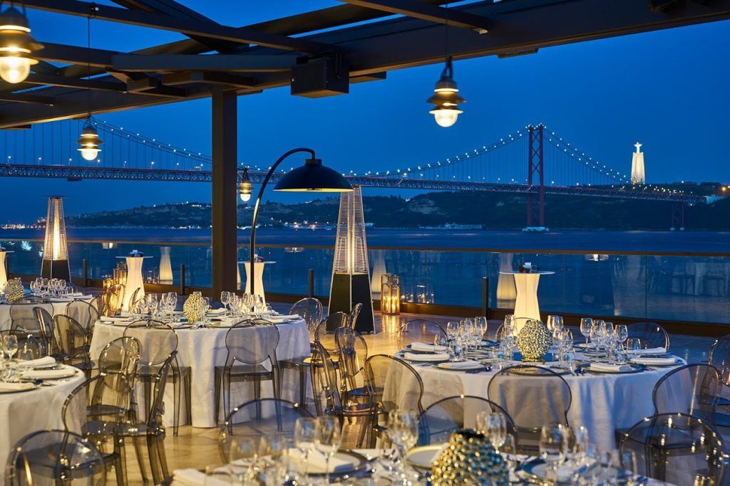Scene of wedding tables set iwth linen table clothes, candle light at SUD Lisboa with view to the Tagus river and teh 25 of April Bridge beyond in the dusk light